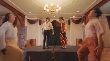 Sahnede dramatik bir dans gösterisi. Beyaz tişörtlü ve jartiyer giymiş bir erkek dansçı ve canlı kırmızı ve siyah elbiseli bir bayan dansçı. Dekorun ahşap aksanlı ve yumuşak ışıklandırmalı kırsal bir atmosferi var..