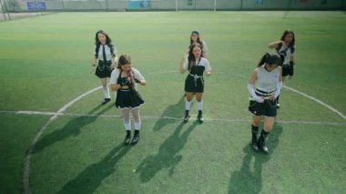 Bir spor sahasında daireler çizen şık kıyafetli altı genç kadın grubu. Kolları çapraz ve başları aşağıda, çimenlerin üzerinde uzun gölgeler bırakarak senkronize bir şekilde dururlar..