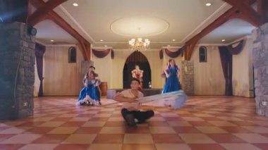 Geniş bir salonda ahşap aksanlı canlı bir dans sahnesi. Çiftler neşeyle dans ederken, bir kişi neşeyle yere düşüyor. Renkli kostümler atmosfere enerji katıyor..