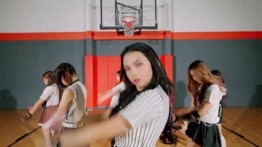 Bir spor salonunda sahne alan dört dansçıdan oluşan bir grup. Etek ve çizgili kıyafetler giyiyorlar. Baş dansçı odak noktasında, enerjik dans figürleri sergiliyor, diğerleri de arka planda onu takip ediyor..