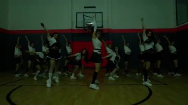 Bir grup dansçı bir spor salonunda koreografi gösterisi yapıyor. Etek ve spor ayakkabılarla uyumlu kıyafetler giyiyorlar, enerjik hareketler sergiliyorlar. Basketbol potası arka planda görünür..