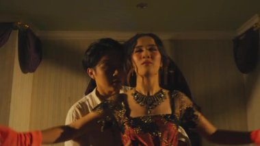 Loş bir odada bir erkek ve bir kadının olduğu dramatik bir sahne. Kadın süslü bir kostüm giyip kolye ve kırmızı eldivenlerle dolaşırken, erkek ona yakın görünüyor ve samimi bir atmosfer yaratıyor..