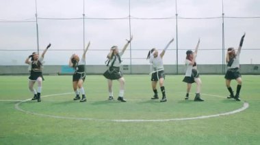 Altı kadın dansçıdan oluşan bir grup spor sahasında koreografili bir gösteri yapıyor. Aynı kıyafetleri giyiyorlar, sportif bir hava katıyorlar, dinamik hareketler ve ifadeler sergiliyorlar. Arka planda yeşil bir çim ve bulutlu bir gökyüzü var..