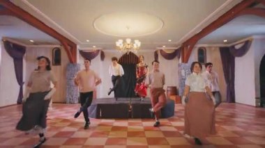 Bir grup dansçı klasik temalı bir ortamda sahnede sahne alıyor. Odada ahşap kirişler, zarif avizeler ve kareli bir zemin bulunuyor. Dansçılar dönemin kostümlerini giyer, canlı dans figürlerini sergilerler..