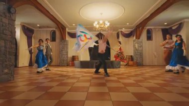 Geniş bir salonda kareli zemini olan canlı bir dans gösterisi. Renkli kostümlü dansçılar sahne alırken, bir dansçı büyük bir pankart taşıyor. Kurulumda taş duvarlar ve zarif dekorlar yer alıyor..