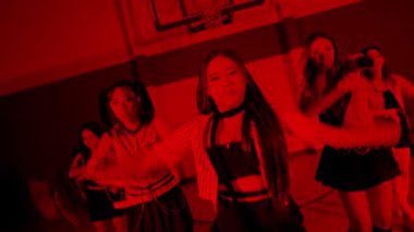 Bir grup kız spor salonunda, kırmızı ışıkla aydınlatılmış, dramatik bir atmosfer yaratıyorlar. Dinamik hareketlerle bir dans ya da performans sergiliyor gibi görünüyorlar..