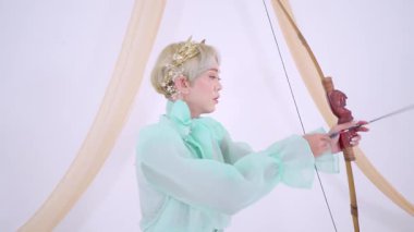 Açık yeşil kıyafetli biri geleneksel bir yayla ok atmaya hazırlanıyor. Arka planda yumuşak perdelerle minimalistik bir ortam yaratıyor..