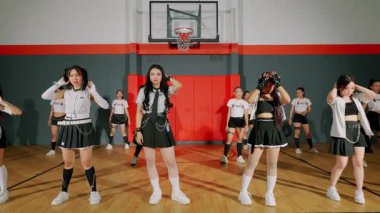 Bir grup dansçı jimnastik salonunda gösteri yapıyor. Etek ve spor ayakkabılarla şık kıyafetler giyiyorlar. Arkaplanda bir basketbol potası ve canlı bir renk şeması yer alıyor..