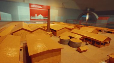 Bir fabrika kompleksinin detaylı mimari modeli, depolama tankları ve baca gibi çeşitli binaları gösteriyor. Modeli ahşaptan yapılmıştır ve arka planda bilgilendirici tabelalar bulunan bir müze ortamında sergilenmektedir..