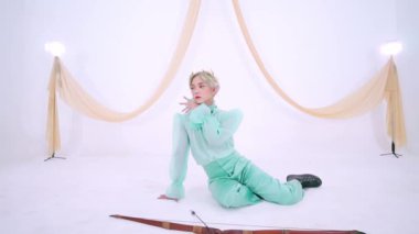 Nane yeşili kıyafetli bir kişi beyaz bir arka planda kumaşla zarif poz verir. Dekor parlak ve minimalistik, öznenin duruşu ve giysisini vurguluyor..