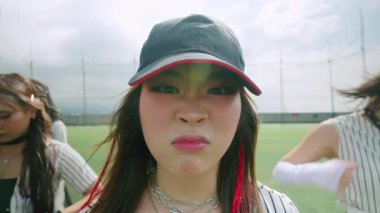 Şapka takmış, şık giyinmiş kameraya gülümseyen genç bir kadının yakın çekimi. Arka planda bulutlu gökyüzü olan bir spor alanı var.