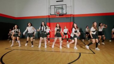 Bir grup dansçı bir spor salonunda koreografi gösterisi yapıyor. Aynı kıyafetleri giymişler, enerjik hareketler sergiliyorlar. Basketbol potası ve kortu arka planda görünüyor..