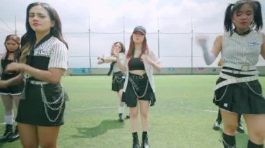 Bir futbol sahasında beş bayan dansçı zincirli ve aksesuarlı, şık siyah-beyaz kıyafetler giyiyorlar. Parlak bir gökyüzünün altında dinamik bir dans pozisyonundalar..