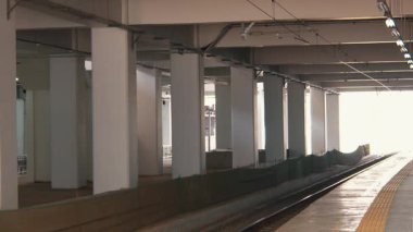 Beton sütunları ve arka planda parlak bir çıkışı olan boş bir tren istasyonu platformu. Bölge yeşil bariyerler ve asgari ışıklandırma ile inşaat halinde görünüyor.