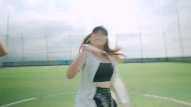 Spor sahasında dinamik bir dans gösterisi, arka planda iki dansçıyla birlikte beyzbol şapkası ve kırbaç bluzu giymiş bir kadın dansçı. Sahne, parlak bir gökyüzü altında enerji ve hareket yakalar..