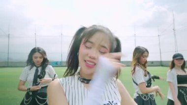 Bir grup genç kadın bir spor sahasında dans gösterisi yapıyor. Ön planda örgülü saçlı, çizgili kıyafetli, gülümseyen bir kız var. Arka plan, yukarıda bulutlu bir gökyüzü ile hareket eden diğer dansçıları gösteriyor..