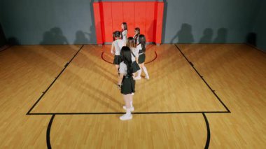 Basketbol sahasında dans eden bir grup genç kadın. Mahkemede bir basketbol potası ve duvarlarda kırmızı aksanlar yer alıyor. Yerdeki gölgeler olay yerine derinlik katar.