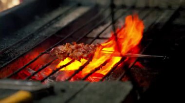 Sıcak kömürlerin üzerinde ızgara bir et şiş, arka planda alevler ve duman yükseliyor, canlı ve iştah açıcı bir sahne yaratıyor.