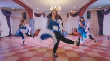 Geniş bir salonda avizeli canlı bir dans gösterisi. Mavi elbiseli dansçılar, hareketlerini sergiliyorlar. Ayarda ahşap kirişler ve zarif dekorlar yer alıyor..