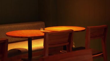 İçinde iki yuvarlak ahşap masa ve ahşap sandalyeler olan rahat bir kafe. Sıcak ışıklandırma, arka planında yumuşak gri bir bank bulunan hoş bir atmosfer yaratıyor..