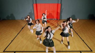 Spor salonundaki bir grup genç bayanın dinamik dans gösterisi. Aynı kıyafetleri giyiyorlar ve arka planda basketbol potası olan senkronize hareketler sergiliyorlar..