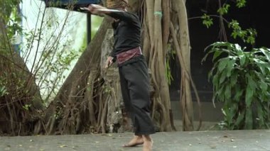 Ağaçlar ve yeşilliklerle çevrili, bereketli bir ortamda dinamik bir hareket yapan bir dövüş sanatçısı. Sahne, aksiyon ve çevikliği, geleneksel kıyafetleri ve odaklanmış ifadeyi yakalıyor..