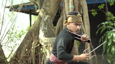 Siyah kıyafetli ve bantlı bir dövüş sanatçısı, ağaçlar ve yeşilliklerle çevrili doğal bir ortamda pençe benzeri silahlarla dramatik bir şekilde poz verir..