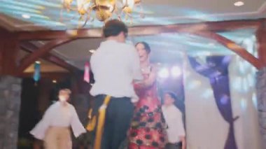 Geleneksel giyinmiş iki çiftin olduğu canlı bir dans sahnesi. Bir çift yakından dans ederken, diğeri arka planda kısmen görülebilir. Dekorun ahşap fıçılar ve örtülü kumaşlarla kırsal bir cazibesi var..