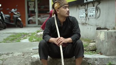 Geleneksel giyinmiş genç bir adam taş basamağa oturmuş, elinde tahta bir sopayla. Siyah bir elbise ve desenli bir başörtüsü takıyor. Uzağa dikkatlice bakıyor. Arka planda park etmiş motosikletli ve binalı bir sokak manzarası var..