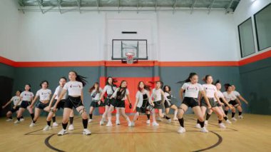 Basketbol salonunda gösteri yapan bir grup dansçı. Aynı kıyafetleri giyiyorlar ve senkronize hareketleri sergiliyorlar. Enerjik bir atmosfer yaratıyorlar. Spor salonunda basketbol potası ve ahşap zemin var..