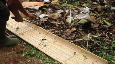 Yeşil çizmeli biri, yapraklar ve çöplerle kaplı pis bir zeminde uzun bir bambu şeridiyle çalışıyor. Sahne, doğal molozlarla dolu bir açık hava ortamını betimliyor..