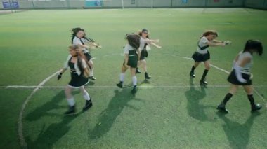 Bir spor sahasında dans eden altı genç kadından oluşan bir grup. Şık kıyafetler giyiyorlar, etek ve üstlerin karışımı, enerjik hareketler sergiliyorlar. Sahne parlak ve canlı, takım çalışmasının ruhunu yansıtıyor.