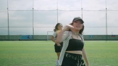 Bir spor sahasında canlı bir dans sahnesi. Üç dansçı şık kıyafetler giyecek. Ön plandaki dansçı hareket halinde, dinamik bir atmosfer yaratıyor. Arkaplan yeşil bir alan ve bulutlu bir gökyüzünü gösteriyor.