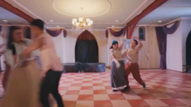 Geniş bir salonda ahşap kirişler ve zarif dekorlarla canlı bir dans gösterisi. Altı dansçı, klasik kıyafetler giymiş, dinamik bir gösteri sergiliyorlar, neşe ve enerji ifade ediyorlar..