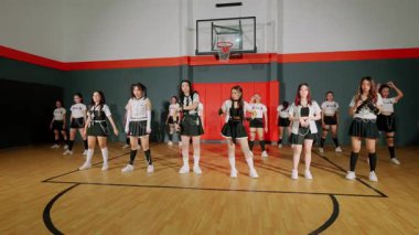 Bir grup genç kadın bir spor salonunda dans gösterisi yapıyor. Koordineli kıyafetler giyiyorlar, enerji ve takım çalışması sergiliyorlar. Arka planda basketbol potası ve saha yer alıyor..