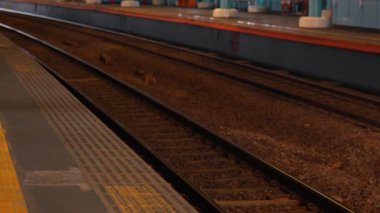 Raylara ve platforma odaklı bir tren istasyonundaki rayların yakın görüntüsü. Sahne boş, parlak, aydınlık bir ortamın altında çakıl ve metal izleri var..