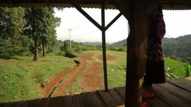 Geleneksel giyinmiş bir kadın ahşap bir platformda duruyor, çatı kirişine tutunuyor. Arka planda yemyeşil tepeler ve görülebilir elektrik hatlarıyla kirli bir patika var..