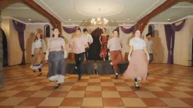 Bir grup dansçı dekore edilmiş bir salonda sahneye çıkıyorlar. Dansçılar geleneksel kostümler giyer, canlı bir dans gösterisi yaparlar. Ayarda ahşap kirişler ve zarif dekorlar yer alıyor..