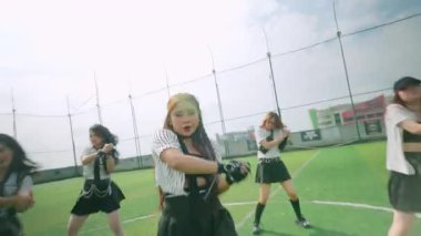 Dinamik bir dans sahnesi. Parlak bir gökyüzünün altındaki çimenli bir alanda birden fazla sanatçı var. Dansçılar hareketli, etkileyici jestlerle enerjik koreografi sergiliyorlar..