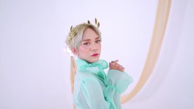 Bir model zarif yeşil bir elbise içinde yüksek yakalı, zarif bir taç ve çiçek aksesuarlarıyla süslenmiş bir şekilde poz verir. Arka plan yumuşak, akışkan kumaşı nötr tonlarda içeriyor.