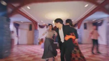 Geniş bir salonda dans sahnesi. Ön planda dans eden bir çift. Kadın canlı bir elbise ve eldiven giyer, erkek ise takım elbise giyer. Diğer dansçılar arka planda hareketli, neşeli bir atmosfer yaratıyorlar..