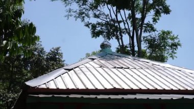 Benzersiz bir tasarımı olan, merkezi havalandırma kapağına sahip metal bir çatının yakın çekim görüntüsü. Çevreleyen ağaçlar açık mavi gökyüzünün altında doğal bir zemin sağlar..