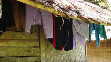 Sazlı bir çatı altında bambu bir yapıda asılı duran giysiler, çeşitli renkler ve stiller sergiliyor. Arka planda geleneksel evlere özgü örülmüş bir duvar yer alıyor..