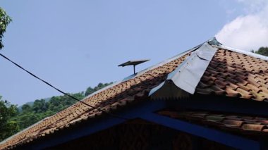 Terracotta fayansları olan ve tepesinde güneş paneli olan bir evin yakın çekim görüntüsü. Gökyüzü birkaç bulutla berrak ve arka planda yeşil tepeler görünüyor..