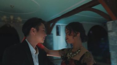 Zarif bir balo salonunda dans eden bir çift, yerde yansıyan renkli ışıklarla. Kadın canlı, çiçekli bir elbise giyer, erkek ise resmi bir takım elbise giyer. Atmosfer romantik ve dramatik..