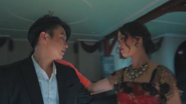 Zarif ve loş bir balo salonunda dans eden bir çift. Kadın, erkek resmi bir takım giyerken, karmaşık desenli kırmızı ve siyah bir elbise giyer. Yumuşak ışıklandırma ve yerde yansımalarla atmosfer romantik..