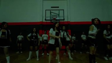 Bir grup dansçı, loş bir spor salonunda, enerjik koreografi sergileyerek performans sergiliyorlar. Dansçılar, hareket ve ifade üzerine odaklanmış şık kıyafetler giyerler..