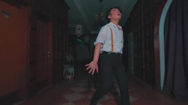 Beyaz gömlekli ve pantolon askısı takmış genç bir adam loş bir odada elinde bir kağıt parçasıyla dikiliyor. Arka planda karanlık perdeler ve dekoratif elementler var. Gizemli bir atmosfer yaratıyor..