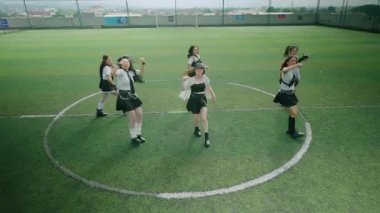 Altı dansçıdan oluşan bir grup yeşil bir spor sahasında koreografili bir gösteri yapıyor. Şık kıyafetler giyiyorlar, siyah ve beyaz renkli karışımlar sergiliyorlar, bazıları eldiven ve aksesuar takıyor. Arkaplanda bulanık bir görünüm var