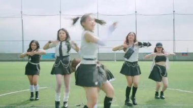 Bir grup genç kadın bir spor sahasında dans gösterisi yapıyor. Şık kıyafetler giymişler. Ön planda bir kadın var. Enerjik bir şekilde kamerayı işaret ediyor. Arkaplanda yeşil bir çim alanı ve bulutlu bir gökyüzü var..